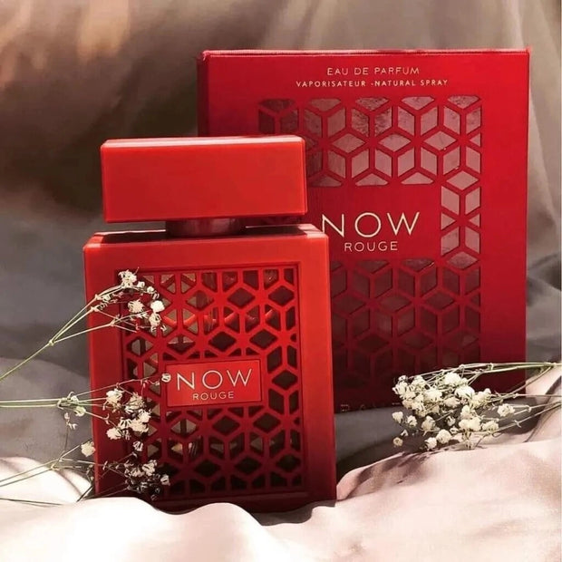 Red 'NOW Rouge' perfume bottle and packaging on a soft surface with flowers.