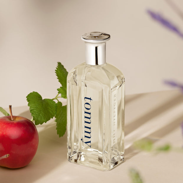 Clear glass perfume bottle with silver cap labeled 'tommy' next to a red apple on a light background