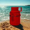 Red thermos with 'Odyssey Artista' branding on a sandy beach with ocean in the background