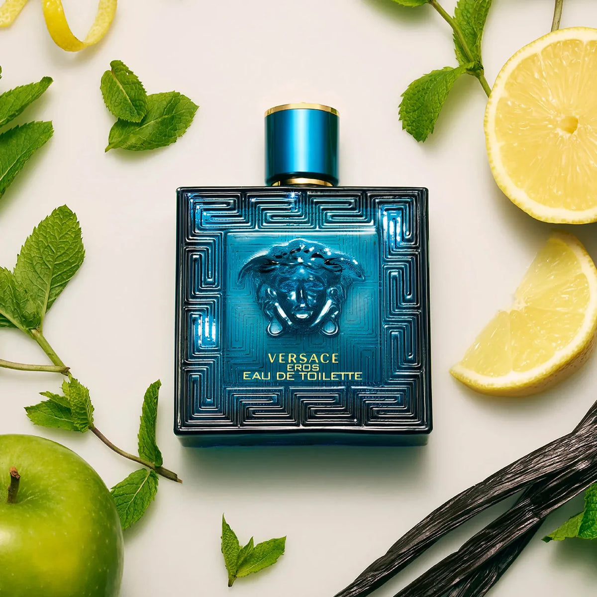 Versace men's perfume bottle surrounded by lemons, apples, and vanilla beans on a light background